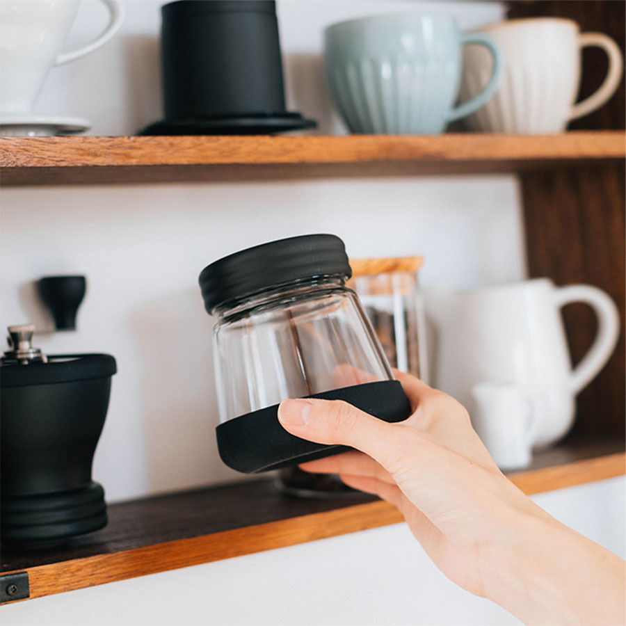 Hand hält Behälter vor Küchenschrank mit Hario Skerton N Handkaffeemühle im Hintergrund.