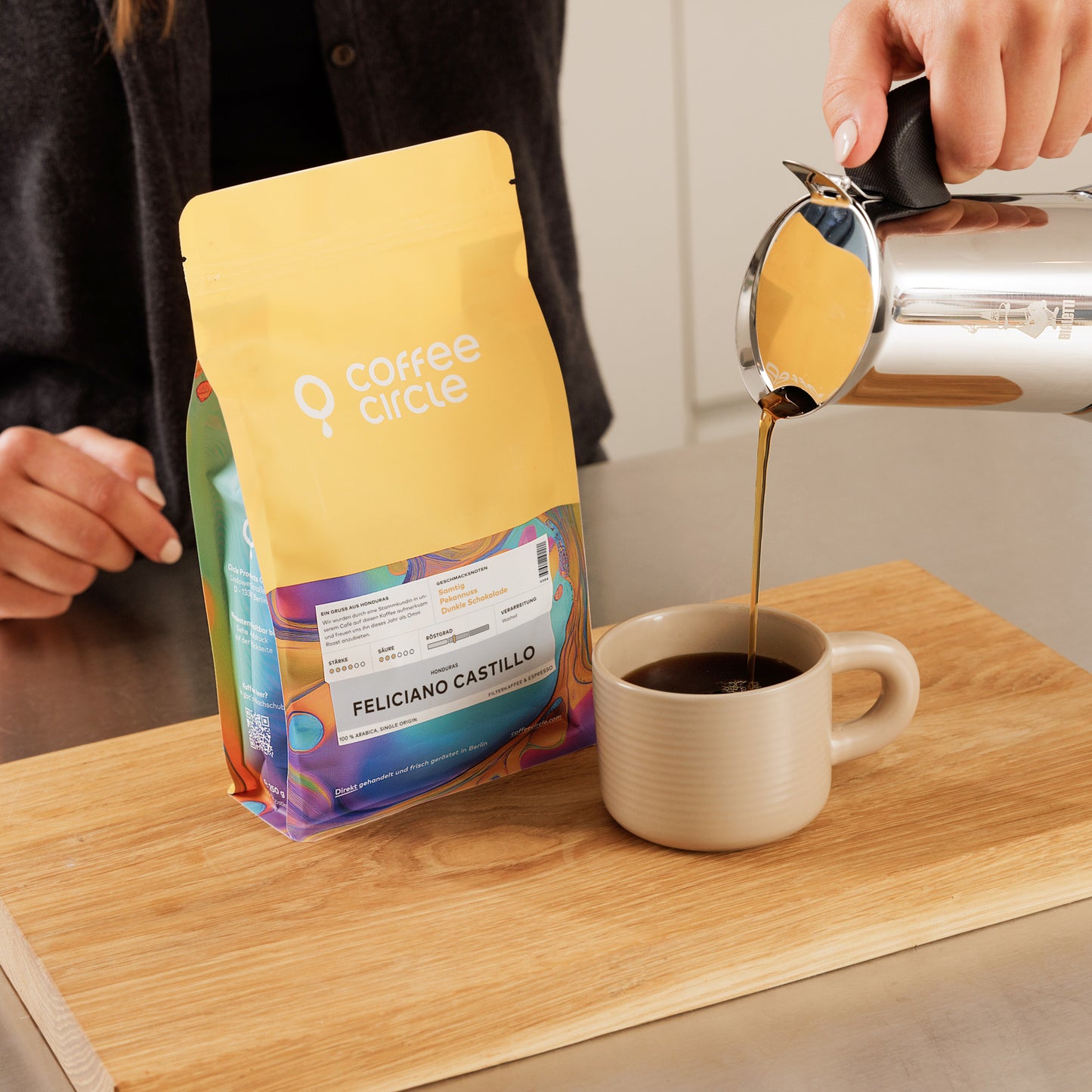 Person gießt Espresso Feliciano Castillo in Tasse neben bunter Coffee Circle Verpackung auf Holztisch.